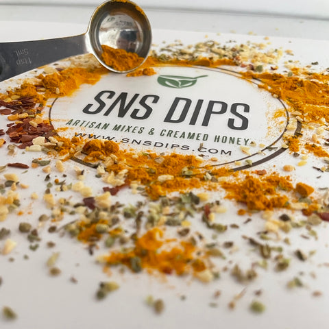 Gourmet dip mix spices and herbs scattered across a white board printed with the SnS Dips Artisan Mixes and Creamed Honey logo