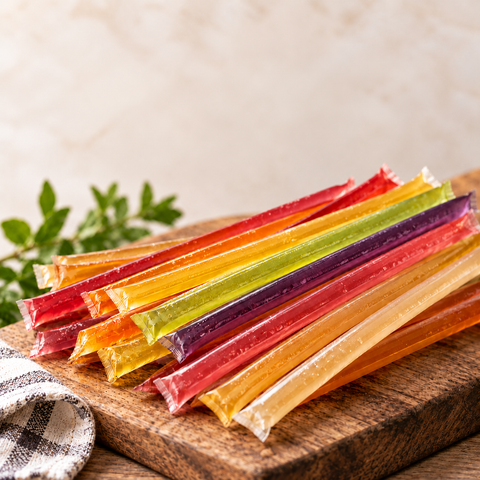 A stack of colorful, individually wrapped honey sticks arranged on a wooden cutting board with a small checkered cloth and green leaves in the background.